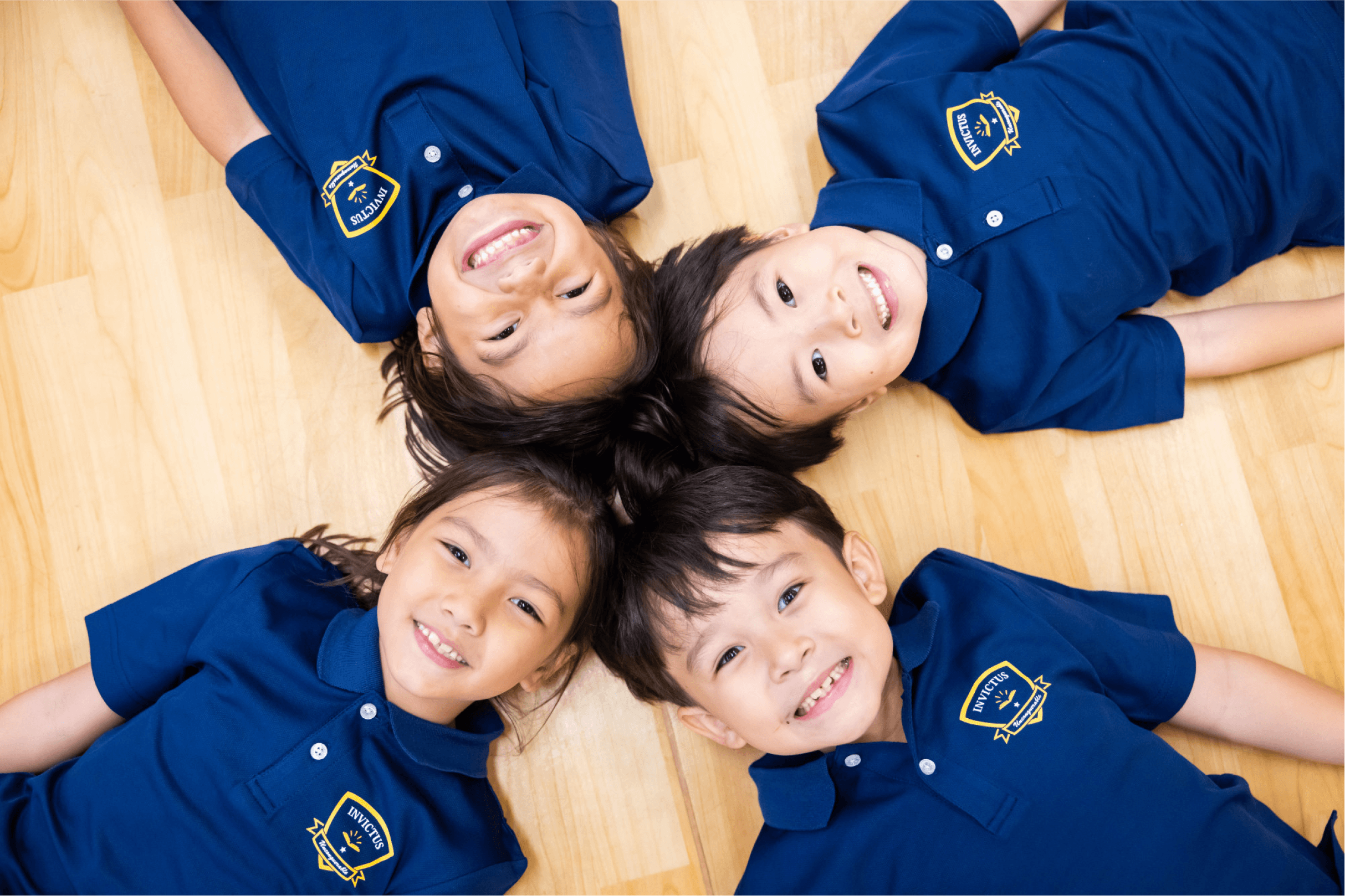 Four kids lying down on the wooden floor and smiling at the camera.